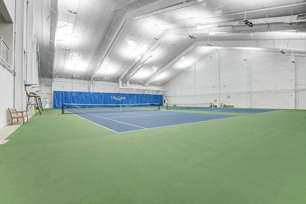 Una cancha de tenis interior con superficie azul y verde, paredes blancas y un techo alto con conductos visibles.