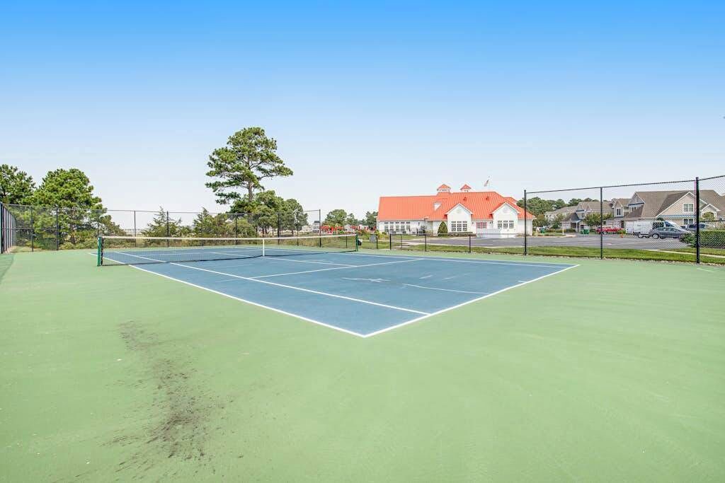 An outdoor tennis court with a net, surrounded by a fence, and a building with a red roof in the background.