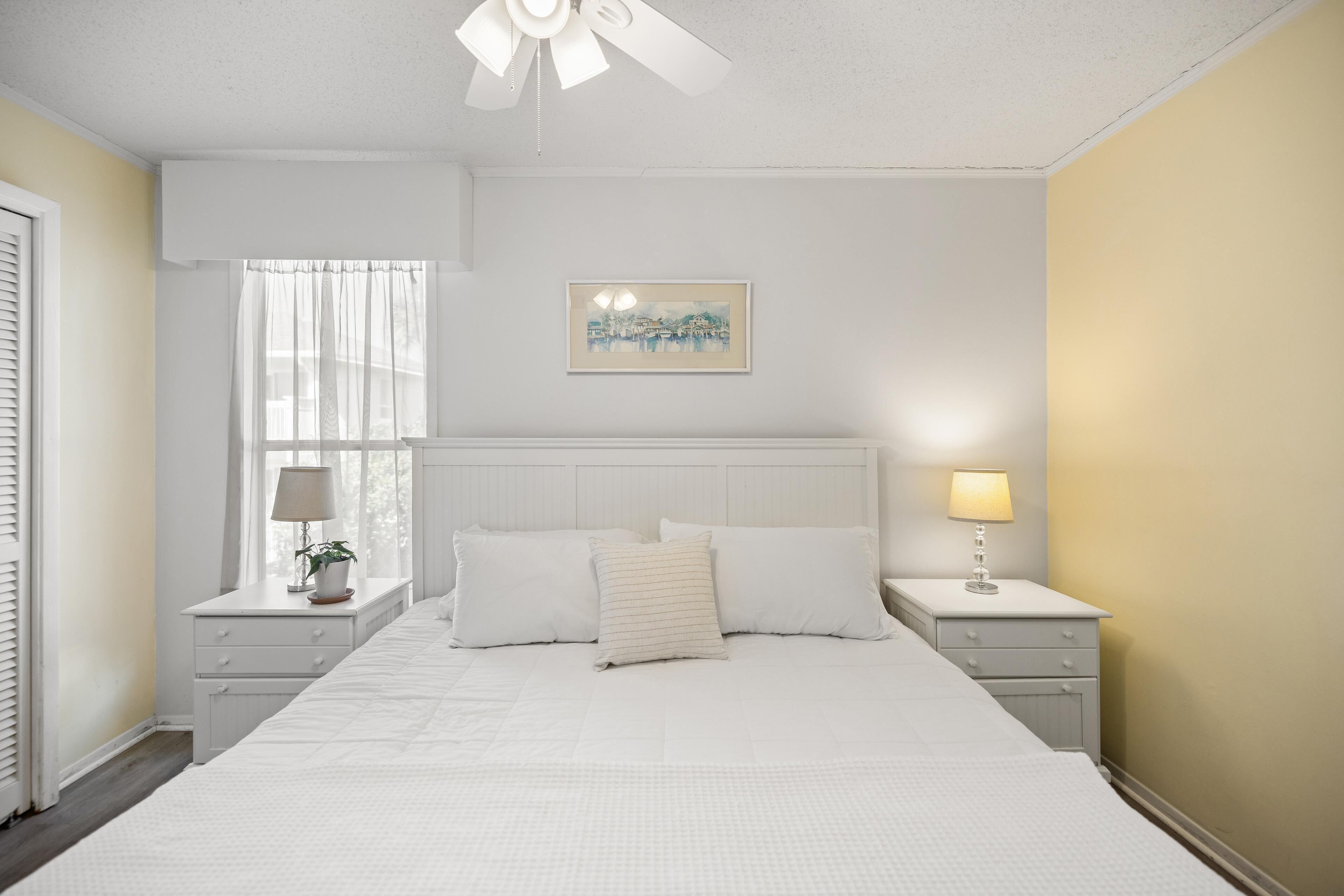 Een slaapkamer met een bed, twee nachtkastjes, een ventilator in het plafond, een raam met gordijnen en een aan de muur hangend schilderij.