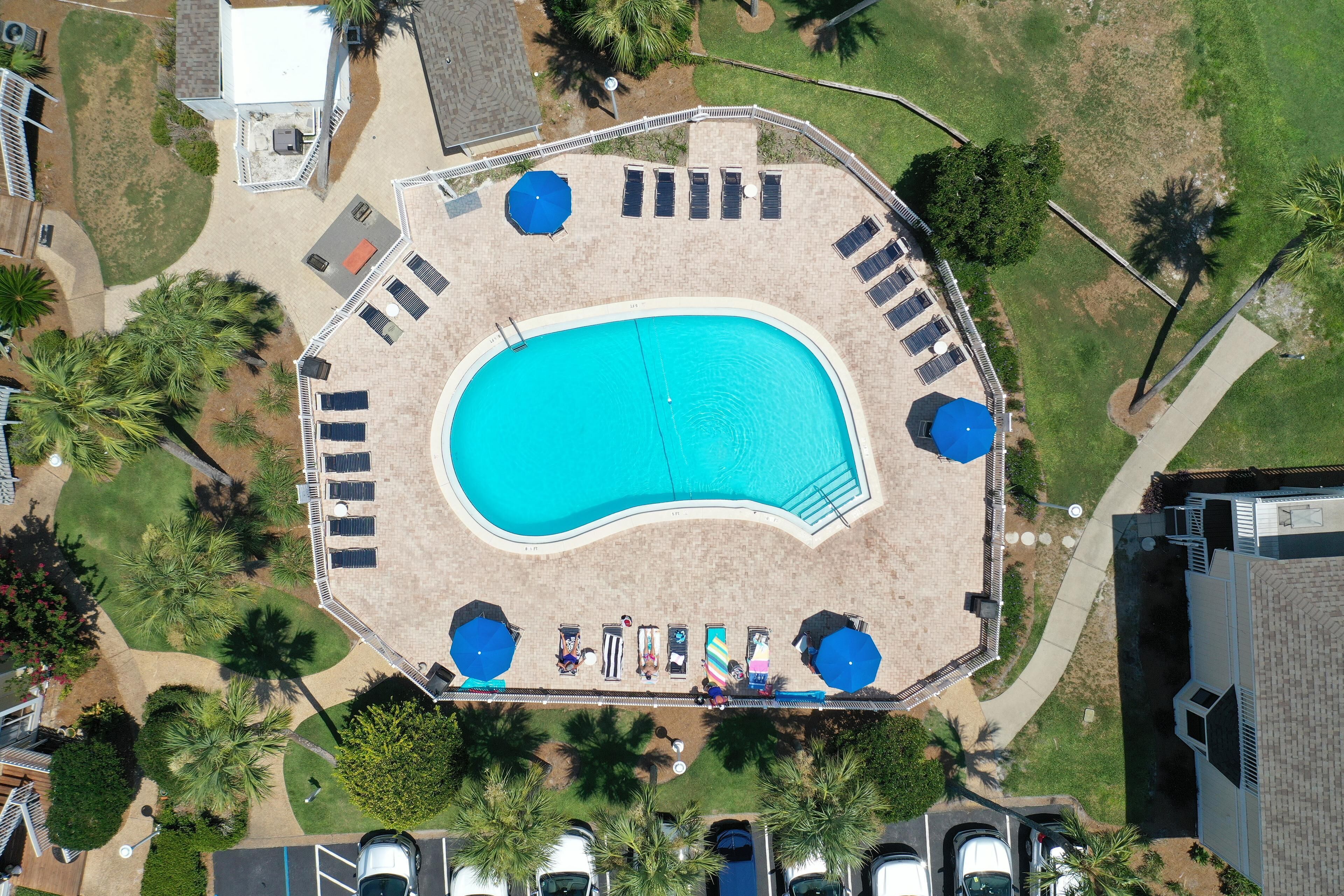 Vista aérea de una zona de piscina con tumbonas, sombrillas y un estacionamiento.