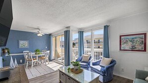 Uma sala de estar com mesa de centro de vidro, sofá azul e um espaço de refeições com cadeiras brancas.