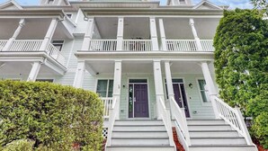 A two-story house with a front porch, a central door, and a staircase leading to the entrance. - Three-Story Bayside Resort Retreat W/ Shared Pool/Hot Tub, Balcony & Central AC (Selbyville)
