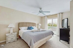 Ein Schlafzimmer mit Bett, Deckenventilator, Nachttischlampen, einem Fernseher und einem Balkon mit Vorhängen.