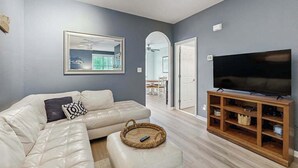 A living room with a sectional sofa, a flat-screen TV on a wooden stand, and a framed picture on the wall. - Three-Story Bayside Resort Retreat W/ Shared Pool/Hot Tub, Balcony & Central AC (Selbyville)