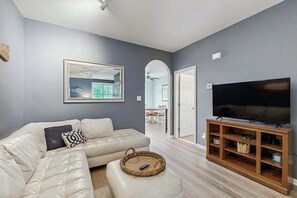 Ein Wohnzimmer mit einer Eckcouch, einem Flachbildfernseher auf einem Holzboden und einem gerahmten Bild an der Wand.
