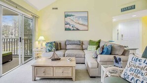 Ein Wohnzimmer mit einer Couch, einem Couchtisch, einem Strandbild und einem Schild mit der Aufschrift 'BEACH HOUSE'.
