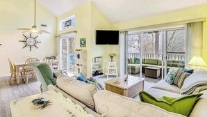 Ein Wohnzimmer mit einer Couch, einem Esstisch mit Stühlen, einem Fernseher und einem Balkon mit Sitzgelegenheit.