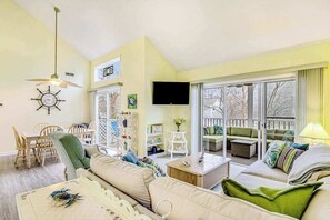 A living room with a sofa, a dining table with chairs, a television, and a balcony with outdoor seating. - Sea Colony Condo w/ Free WiFi, Central AC, Shared Pools, & Hot Tubs (Bethany Beach)