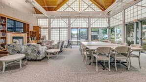 A spacious room with a high ceiling, large windows, a fireplace, and a dining area with chairs and tables. - Bear Trap Dunes Townhouse Near Bethany Beach w/Shared Pool, Free WiFi, W/D (Ocean View)