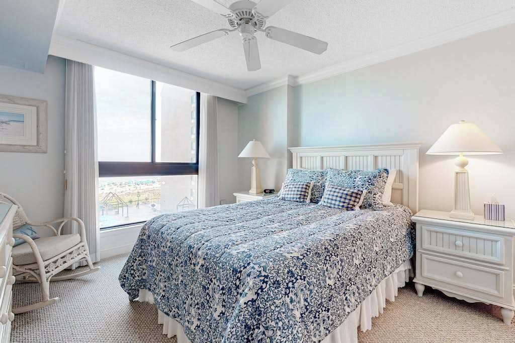 A bedroom with a bed, two bedside tables, a chair, a ceiling fan, and a window.