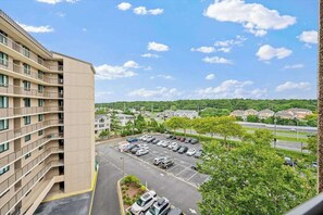 A multi-story building with a parking lot in front, surrounded by greenery and a clear sky. - Sea Colony Ocean condo with shared pools, hot tubs, beach access & balcony (Bethany Beach)