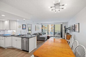 Eine moderne Küche mit einer zentralen Kochinsel, ein Essbereich mit Holztisch und -stühlen sowie ein Wohnbereich mit Sofa und Fernseher.