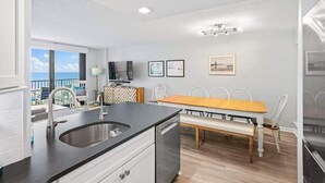 Una cocina moderna con lavavajillas incorporado, un comedor con mesa de madera y sillas blancas, y un balcón con vistas al mar.