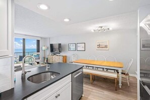 Eine moderne Küche mit eingebautem Geschirrspüler, ein Essbereich mit Holztisch und weißen Stühlen sowie ein Balkon mit Meerblick.