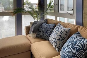 Ein Sofa mit gemusterten Kissen und einer Pflanze, ein Fenster mit Blick auf Wasser und Gebäude sowie eine Tür mit Glasfenster.