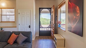 Un couloir avec une porte donnant sur un balcon, un canapé orné de coussins à motifs, une petite table et un grand tableau encadré au mur.