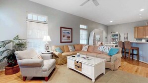 Uma sala de estar com sofá, poltronas, mesa de centro, área de jantar com mesa e cadeiras, e área de cozinha com armários e bancada.
