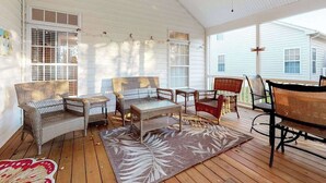 Varanda coberta com móveis de vime, tapete estampado e vista para uma casa branca.