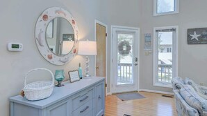 A room with a white dresser, a round mirror with a floral frame, a basket, a lamp, and a blue glass. There are two doors, one with a window and another with a view of a house. - Dog-friendly home close to the beach w/balcony, fireplace, grill, AC (Bethany Beach)