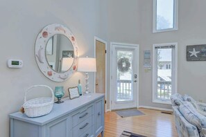 A room with a white dresser, a round mirror with a floral frame, a basket, a lamp, and a blue glass. There are two doors, one with a window and another with a view of a house. - Dog-friendly home close to the beach w/balcony, fireplace, grill, AC (Bethany Beach)