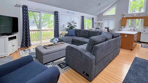 A modern living room with a sectional sofa, a flat-screen TV, and a kitchen area with a window. - Dog-friendly home close to the beach w/balcony, fireplace, grill, AC (Bethany Beach)
