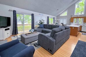 A modern living room with a sectional sofa, a flat-screen TV, and a kitchen area with a window. - Dog-friendly home close to the beach w/balcony, fireplace, grill, AC (Bethany Beach)