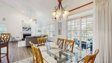 A spacious living room with a glass dining table, wooden chairs, a fireplace, and large windows.