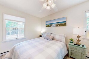 Une chambre à coucher avec un lit, une table de chevet, un ventilateur de plafond, une fenêtre et un tableau accroché au mur.