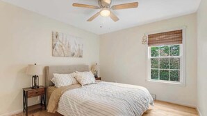 Un dormitorio con cama, mesita de noche, ventilador de techo, ventana con persianas y una obra de arte colgada en la pared.