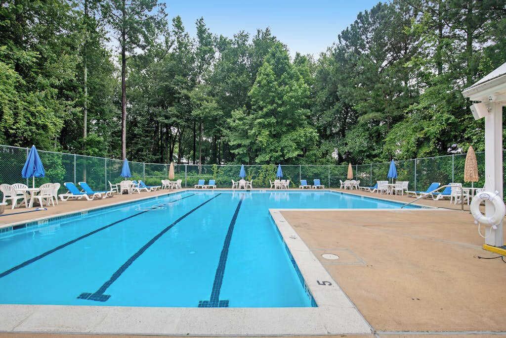 Ein Schwimmbecken mit mehreren Bahnen, umgeben von Liegestühlen und Sonnenschirmen, mit einem Zaun und Bäumen im Hintergrund.