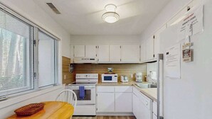 Cocina compacta con armarios blancos, microondas, horno y refrigerador. Hay una mesa de madera y una silla.