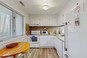Eine kleine Küche mit weißen Schränken, einer Mikrowelle, einem Backofen und einem Kühlschrank. Ein Holztisch und ein Stuhl sind ebenfalls vorhanden.