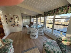 A room with a wooden floor, white wicker chairs, a glass table, and a view of a body of water through large windows. - South Bethany bay-view home with dock, deck, kayaks, & bikes (Bethany Beach)