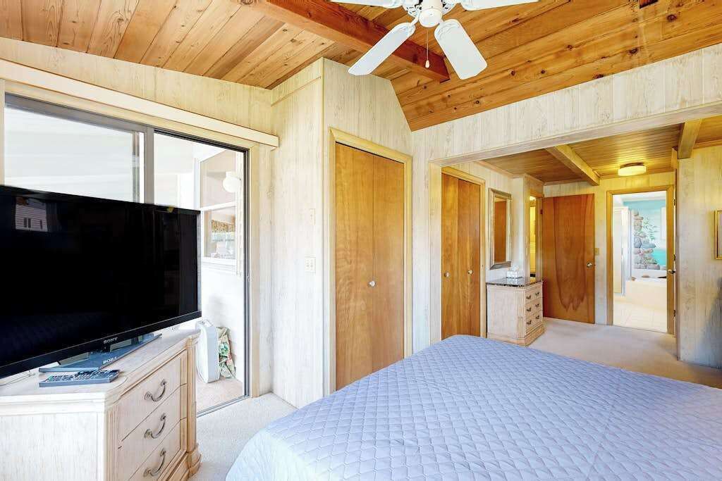 Un dormitorio con cama, televisión, paredes de madera, ventilador de techo y vista por la ventana.