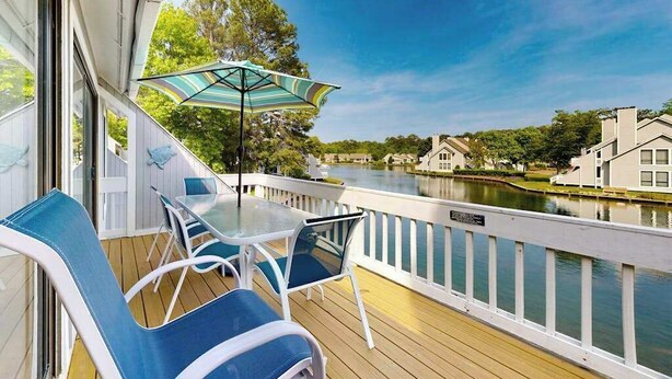 Eine Terrasse mit Tisch und Stühlen, einem gestreiften Sonnenschirm und Blick auf einen Fluss sowie Gebäude.