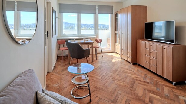 Un soggiorno moderno con pavimento a spina di pesce, una zona pranzo con tavolo e sedie, un televisore su un mobile in legno e un'ampia finestra con vista sulla città.