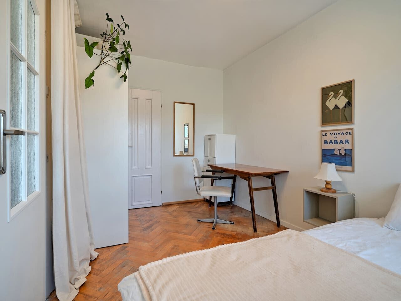Habitación de hotel con cama, escritorio, silla y una mesita de noche con lámpara. Se ve una ventana con cortinas, una puerta y un cartel en la pared.