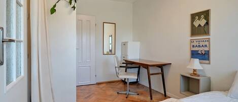 Habitación de hotel con cama, escritorio, silla y una mesita de noche con lámpara. Se ve una ventana con cortinas, una puerta y un cartel en la pared.