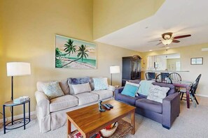 Un salon comprenant deux canapés, une table basse, un tableau représentant une plage, un ventilateur de plafond et un coin repas avec des chaises et une table.