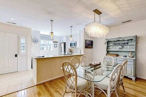 A dining area with a glass table and wicker chairs, a kitchen with white cabinets, and a chandelier.