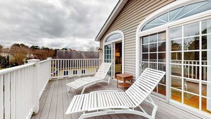 En overdækket terrasse med to hvide liggestole og et lille bord, et hvidt hegn og en stor glasdør, der fører til et overdækket område.
