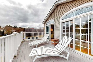 Eine überdachte Terrasse mit zwei weißen Liegestühlen und einem kleinen Tisch, einem weißen Zaun und einer großen Glasdoor, die zu einem überdachten Bereich führt.