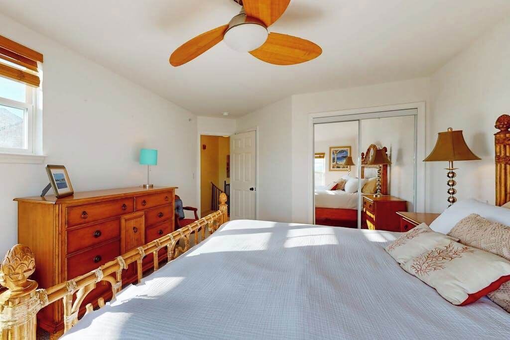 Un dormitorio con cama, cabecera de madera, ventilador de techo, cómoda, lámpara y una puerta que da a otra habitación.