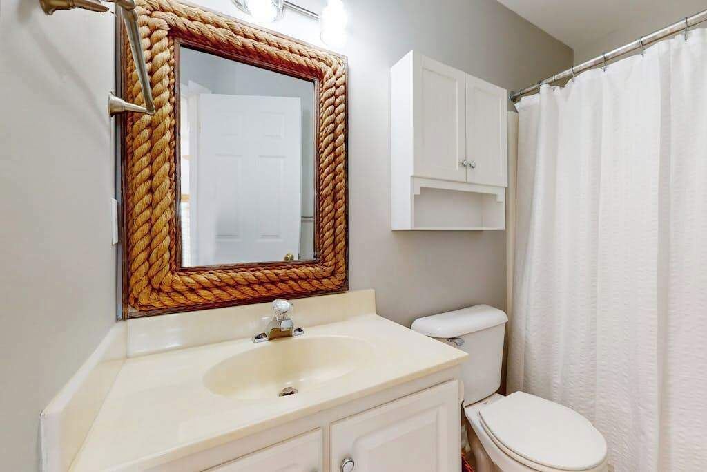 A bathroom with a white sink, a toilet, a mirror with a rope frame, and a white shower curtain.