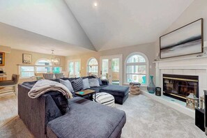 A spacious living room with a fireplace, large windows, and a modern chandelier. - Spacious Salt Pond home with private deck & pool/hot tub/tennis access (Bethany Beach)