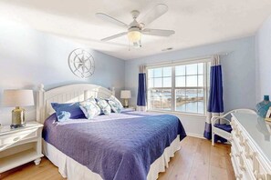 Ein Schlafzimmer mit einem Bett, einem Nachttisch, einem Schreibtisch, einem Stuhl, einem Deckenventilator, einem Fenster mit Vorhängen und einer Wanduhr.