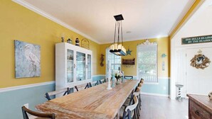 Une salle à manger avec une longue table en bois, des chaises, un buffet blanc aux portes vitrées et un luminaire suspendu.