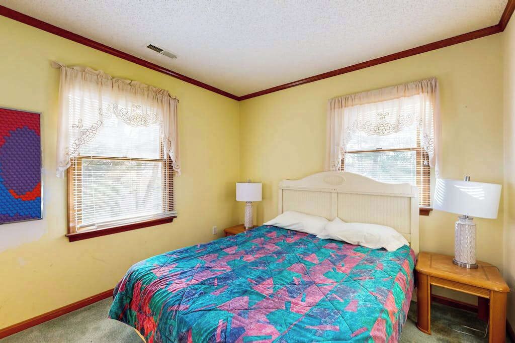 A bedroom with a bed, two bedside tables, a window with curtains, and a wall-mounted lamp.