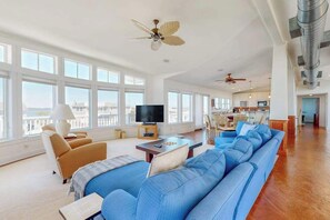 Un salón amplio con un sofá azul grande, una televisión y un comedor, con una cocina al fondo.