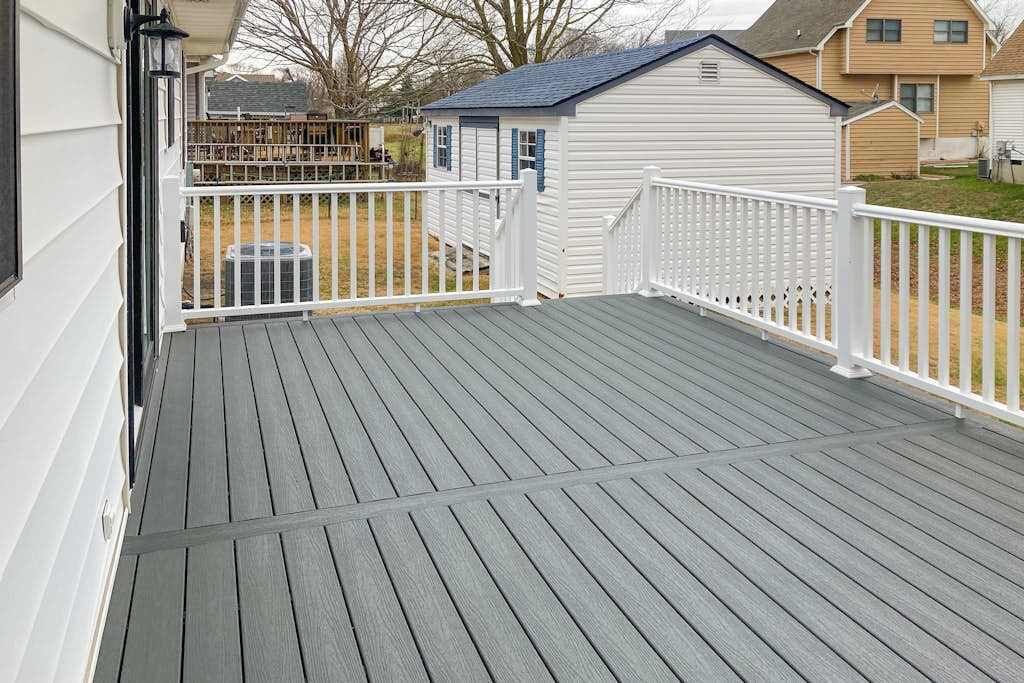 Eine Holzterrasse mit weißen Geländern, eine kleine Gartenhütte und ein Haus im Hintergrund.